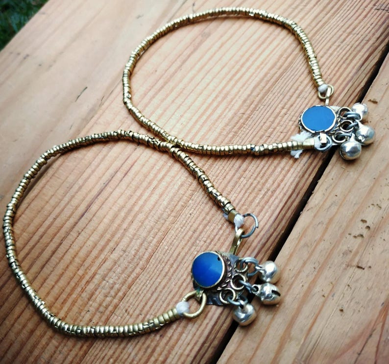 Two gold-tone beaded anklets with blue cabochons and hanging bell charms displayed diagonally on wooden planks.