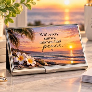 May include: A rectangular silver metal business card holder featuring a printed image of a tropical beach at sunset with palm leaves and white plumeria flowers. The text on the holder reads, With every sunset, may you find peace.