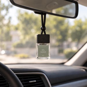 Peut inclure: Un petit flacon diffuseur de parfum pour voiture en verre transparent avec un bouchon en bois noir et un cordon noir suspendu au rétroviseur. L'étiquette vert sauge porte le texte blanc MAISON L'APOGEE SEL SAUGE et 8ml en bas.
