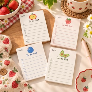 May include: Four rectangular to-do list notepads with white paper and lined pages. Each notepad features a unique fruit illustration at the top: a red apple, a red strawberry, a blue blueberry, and green avocado halves, with the text To do list.