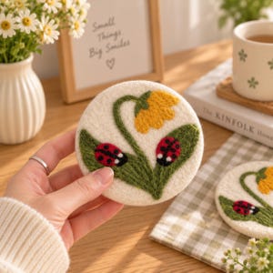 Puede incluir: Un posavasos redondo de fieltro color blanco roto con un diseño bordado de una flor amarilla con tallos y hojas verdes, acentuado por dos pequeñas mariquitas rojas con puntos negros. La textura es suave y fibrosa, con bordados detallados.