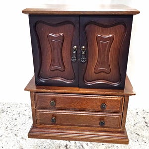 May include: A wooden jewelry armoire featuring a dark brown finish on the upper cabinet doors and a lighter brown finish on the two lower drawers. The cabinet doors have two small metal drop pulls, while the drawers feature four small round knobs.