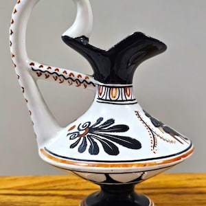 May include: A ceramic pitcher featuring a white base with black and orange painted details. The vessel has a high, curved handle, a flared spout, and a pedestal base. The body is decorated with black floral motifs and geometric patterns.