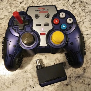 Peut inclure: Une manette de jeu sans fil Nyko Air Flo violette avec un joystick rouge, un stick analogique jaune et des boutons A, B, X, Y multicolores. La manette possède un panneau central argenté avec des réglages de vitesse et un voyant d'alimentation.
