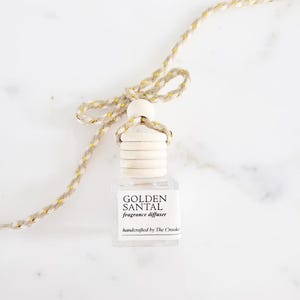 May include: A clear glass fragrance diffuser with a wooden cap and a decorative rope. The label reads "GOLDEN SANTAL fragrance diffuser" and "handcrafted by The Crooks". The rope is beige with gold accents.