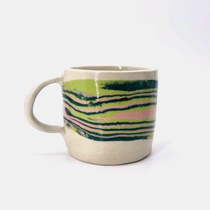 Peut inclure: Une tasse en céramique avec une base blanc cassé lisse et une anse sur le côté gauche. L'extérieur présente un motif marbré horizontal composé de bandes superposées vert foncé, vert lime et rose pâle qui entourent le corps cylindrique.