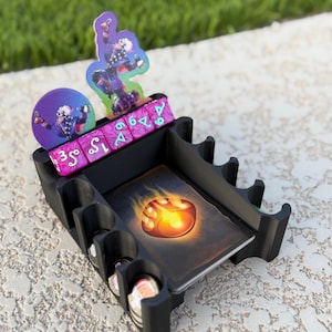 May include: A black plastic tabletop game organizer featuring a central card slot, side compartments for tokens, and a back row for dice. It includes a purple die with white symbols, a circular character token, and a character standee.