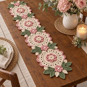 Può includere: Un lungo runner da tavola rettangolare all'uncinetto caratterizzato da un motivo ripetuto di centrini circolari color crema, ciascuno centrato con un fiore rosa. Il runner è decorato con piccoli fiori rosa e foglie all'uncinetto verde scuro.