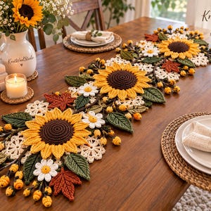 Può includere: Un runner da tavola all'uncinetto con grandi girasoli gialli dal centro marrone scuro, margherite bianche, foglie verdi, foglie d'acero arancioni e piccole bacche gialle, disposto su un tavolo di legno con una brocca bianca.