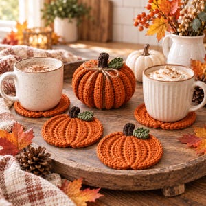 May include: A set of three orange crochet pumpkin coasters with dark brown stems and green leaves, displayed on a wooden tray alongside two ceramic mugs filled with a warm beverage, a larger crochet pumpkin, and autumn-themed decorations.