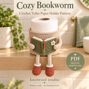 Puede incluir: Un portarrollos de papel higiénico de ganchillo beige con un personaje que sostiene un pequeño libro verde con una flor blanca. El personaje tiene piernas colgantes con zapatos color óxido. Texto: Cozy Bookworm Crochet Toilet Paper Holder Pattern.