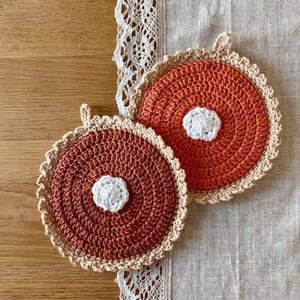 May include: Two circular, hand-crocheted coasters featuring a deep rust-colored center, a small white floral-like accent in the middle, and a scalloped beige border. Each coaster includes a small loop at the top for hanging or storage purposes.