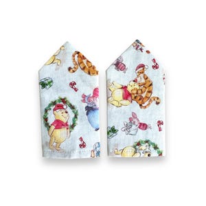 May include: Two light blue fabric napkins with a pointed top edge, featuring a repeating watercolour print of Winnie the Pooh characters including Tigger, Piglet, and Eeyore, alongside Christmas wreaths, gift boxes, and red candy canes.