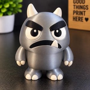May include: A gray monster figurine with a smooth finish, featuring two small white horns, thick black eyebrows, large white eyes with black pupils, a wide black mouth, and a single white tooth. The creature has small white claws on its hands and feet.