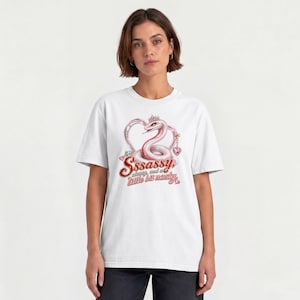 May include: A white short-sleeved t-shirt featuring a graphic of a pink snake wearing a crown, positioned inside a heart shape. Below the snake, the text reads Sssassy, classy, and a little bit nasty in a red and pink stylised cursive font.