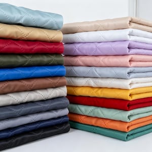 以下が含まれることがあります： ダイヤモンド型のキルティングパターンが施された、折り畳まれた布の正方形の山。ライトブルー、ベージュ、赤、オリーブグリーン、ロイヤルブルー、茶色、グレー、ネイビー、黒、白、ラベンダー、ピンク、黄色、オレンジなど、さまざまな無地のカラーが含まれています。