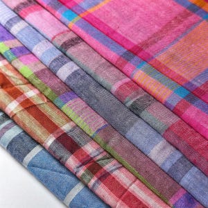 May include: A collection of folded fabric swatches featuring a variety of plaid and checkered patterns. The textiles display a vibrant color palette including shades of pink, blue, orange, green, purple, and red, with visible woven textures.