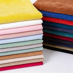 May include: A stack of folded corduroy fabric in a wide variety of colours including yellow, white, pink, light blue, sage green, tan, and red. The fabric features a distinct vertical ribbed texture throughout the entire collection of textile pieces.