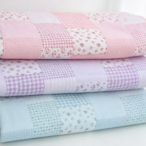 May include: A stack of three folded quilts featuring a patchwork design. Each quilt displays a repeating pattern of squares with alternating floral prints, gingham checks, and solid colors in soft shades of pink, purple, and light blue.