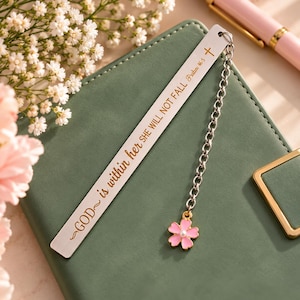 May include: A silver-toned metal bookmark engraved with the text GOD is within her SHE WILL NOT FALL Psalm 46:5 and a small cross symbol. A silver chain is attached to the top, featuring a pink five-petaled flower charm with a white center.