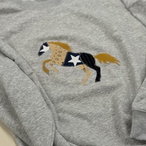 Puede incluir: Una sudadera gris jaspeado de manga larga con una aplicación central de un caballo. El caballo está confeccionado con varios parches de tela en color beige, negro y azul marino, con una estrella blanca en el flanco y en la frente.