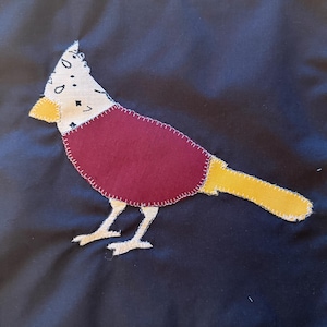 May include: A dark navy fabric background featuring a bird applique. The bird has a white head with black star patterns, a maroon body, a yellow beak, a yellow tail, and white legs, all attached with visible white blanket stitching around the edges.