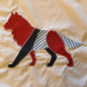 Op de afbeelding: Een stoffen applicatie in de vorm van een wolf of hond, genaaid op een witte achtergrond. Het dier is gemaakt van rode stof met witte stippen, een effen donkerblauwe stof en een lichtblauw met wit geruit vichy-patroon met zichtbare stiksels.