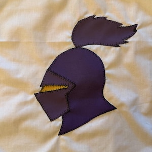 Peut inclure: Un carré de tissu blanc orné d'une applique en forme de casque de chevalier violet avec une visière jaune et un panache violet. Le casque et le panache sont fixés au fond blanc par des points de feston noirs visibles sur les bords.