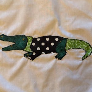 May include: A white fabric background featuring a stitched applique of an alligator. The alligator has a dark green head, a black torso with white polka dots, and a light green tail with a white floral pattern, all outlined with dark stitching.