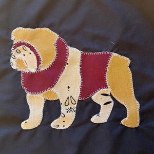 Peut inclure: Une applique en tissu représentant la silhouette d'un bouledogue cousue sur un fond noir. Le chien présente des sections alternées de tissu jaune moutarde et bordeaux, avec des détails en fil blanc et des motifs noirs sur les pattes.