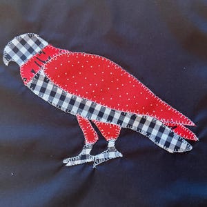 Puede incluir: Una aplicación de tela con forma de pájaro cosida sobre un fondo negro liso. El pájaro presenta un cuerpo rojo con lunares blancos, mientras que la cabeza, las alas y las patas están hechas de tela de cuadros blanco y negro con costuras blancas visibles.