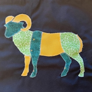 Pode incluir: Uma aplicação de tecido em forma de carneiro sobre um fundo preto, com cabeça verde-escura, chifres amarelos e um corpo composto por retalhos alternados de tecido verde-claro com estampa floral, amarelo sólido e verde-escuro com pequenas estrelas douradas.