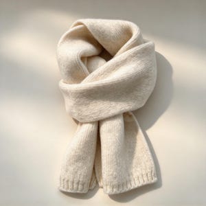 May include: A soft, cream-colored knitted scarf featuring a ribbed texture throughout the length. The scarf is looped and folded over itself, displaying a thick, plush knit construction with finished edges at the bottom ends.