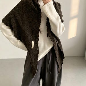 May include: A dark brown triangular crochet shawl with a scalloped edge and a small rectangular white fabric label featuring black text. The shawl is worn over a white long-sleeved ribbed sweater and loose-fitting dark gray trousers.