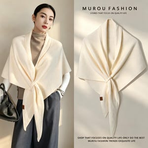 May include: A cream-coloured triangular knit shawl featuring a wide collar and a knotted front closure. The garment includes a small brown rectangular brand tag on the lower edge. The text MUROU FASHION STORES THAT FOCUS ON QUALITY LIFE is visible.