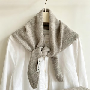 May include: A light beige knit scarf tied in a knot over a crisp white button-down shirt. The scarf features a small white fabric tag with the black text Modeng Mi attached to one end. The shirt has a classic collar and a vertical button placket.