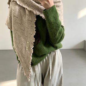 May include: A beige triangular knit scarf with a delicate openwork pattern and a ruffled edge, featuring a small rectangular fabric label with text. The scarf is worn over a fuzzy green long-sleeve sweater and light-colored trousers.