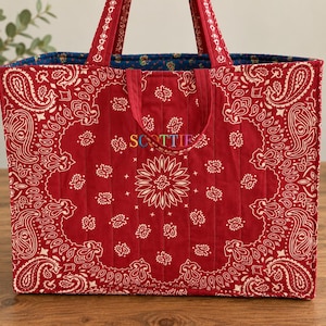 Può includere: Una borsa tote trapuntata con un motivo bandana paisley rosso e bianco. La borsa ha due manici e il nome SCOTTIE ricamato in lettere multicolori sul davanti. La fodera interna è realizzata in tessuto floreale blu scuro.