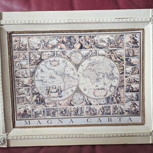 Puede incluir: Un marco rectangular de madera color blanco hueso que contiene una impresión de un mapa del mundo de estilo antiguo. El mapa presenta dos hemisferios circulares rodeados de pequeños paneles ilustrados. El texto MAGNA CARTA aparece abajo.