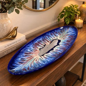 May include: An elongated oval ceramic tray featuring a vibrant, multi-coloured glaze with deep blue, red, and white radial patterns. The centre of the tray has a smooth, irregular, dark grey cutout section that contrasts with the textured, colourful rim.