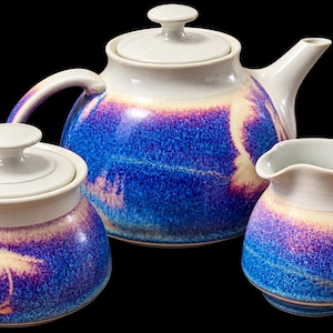 May include: A ceramic tea set consisting of a teapot with a lid, a sugar bowl with a lid, and a creamer pitcher. Each piece features a vibrant blue and purple mottled glaze with cream-colored accents on the rims, handles, and lids.