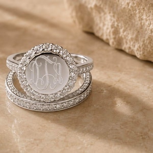 May include: A silver-toned ring set featuring a circular centerpiece with an engraved monogram surrounded by a halo of small clear stones. The band and the halo setting are encrusted with additional clear stones, creating a sparkling effect.
