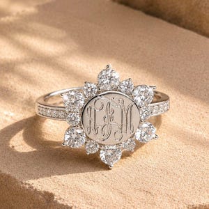 May include: A silver-toned ring featuring a central circular disc engraved with a three-letter monogram. The disc is surrounded by a halo of ten round, clear, faceted stones arranged in a floral pattern, with additional small stones set along the band.