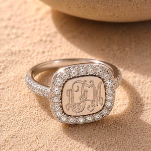 May include: A silver-toned ring featuring a square-shaped face with rounded corners, engraved with the monogram letters LFM. The face is surrounded by a halo of small, clear, sparkling stones, and the band is also encrusted with similar stones.