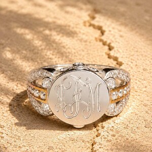 Puede incluir: Un anillo de tono plateado con una cara circular grabada con las letras del monograma LJM. La banda está acentuada con dos filas de pequeñas piedras claras y una franja central dorada, con piedras claras adicionales en los lados del anillo.