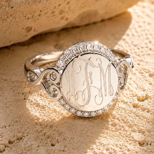 May include: A silver-toned ring featuring a circular face engraved with the monogram letters JFM. The center disc is surrounded by a halo of small, clear stones, and the band displays intricate, openwork scroll designs on both sides.