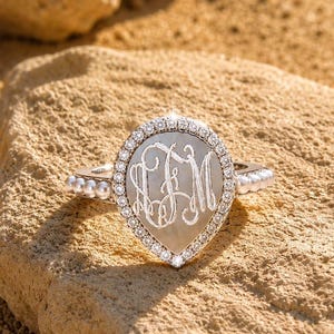 May include: A silver-toned ring featuring a pear-shaped centerpiece with an engraved monogram. The center is surrounded by a halo of small clear stones, and the band is accented with a row of small white pearls on each side.