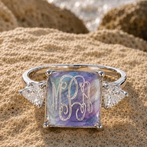 May include: A silver ring featuring a square, iridescent, multi-colored stone engraved with a three-letter monogram. The center stone is flanked by two clear, triangular-cut stones set in a polished silver band with four prongs.