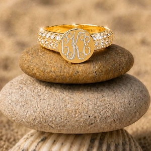May include: A gold ring featuring a circular face with an engraved monogram of the letters C, K, and E in a script font. The band of the ring is encrusted with multiple small, clear, round-cut stones that extend around the entire circumference.