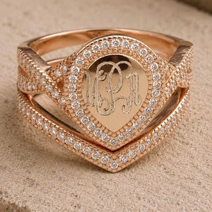 May include: A rose gold ring featuring a pear-shaped center plate engraved with the letters MPJ. The plate is surrounded by a halo of small clear stones. The band consists of two intersecting, V-shaped segments encrusted with clear stones.